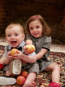 Two children, a girl and a boy, sitting on a rug and holding apples and a bottle of honey