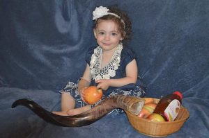 little girl named Kira Berch poses on a blue background wearing a blue dress and holding an apple, next to a basked of apples, a bottle of honey and a shofar